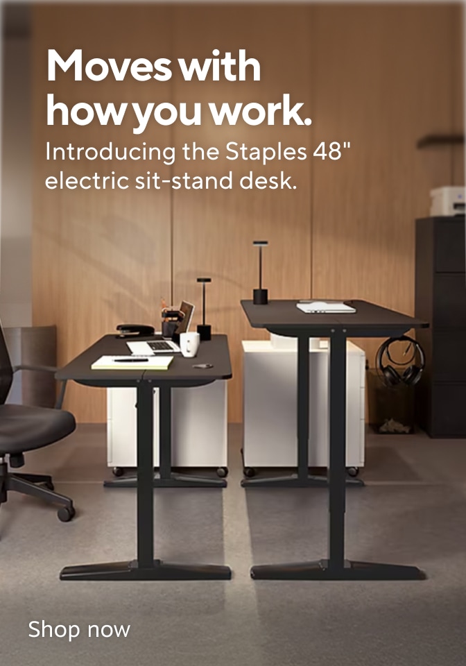 Moves with how you work. Introducing the Staples 48 inch electric sit-stand desk. Image shows two black height-adjustable desks at different heights in a modern office setting with a chair, notebooks, a coffee mug, and desk accessories against a wood-paneled wall. Shop now.