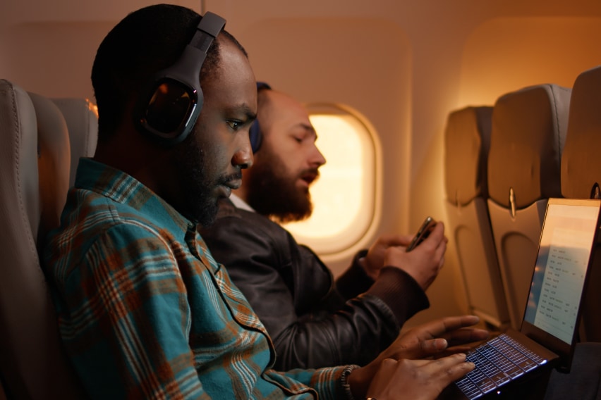 Working while traveling? With a few essential items, you can turn your next flight into a productive work session. Man on a plan working on a laptop with a man next to him on his phone. https://www.staples.com/stores/articles/working/working-while-traveling-5-must-haves-for-your-next-flight.html